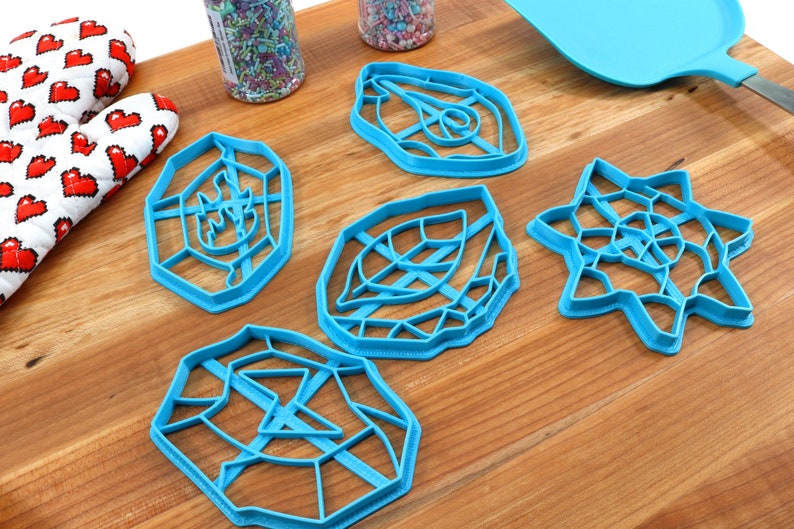 May include: Five blue plastic cookie cutters on a wooden surface. The cutters are shaped like a leaf, a star, a geometric design, and two different geometric shapes.