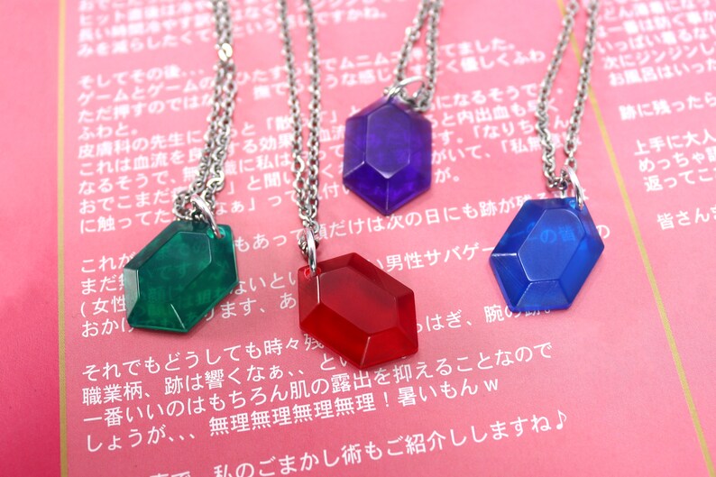 May include: Four necklaces with different colored gem-shaped pendants. The pendants are red, blue, green, and purple. The necklaces are silver chains.