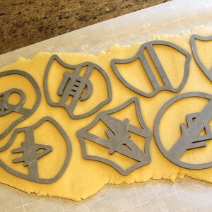 May include: A sheet of golden cookie dough with various cookie cutters pressed into it. The cutters are shaped like shields, circles, and other geometric shapes.