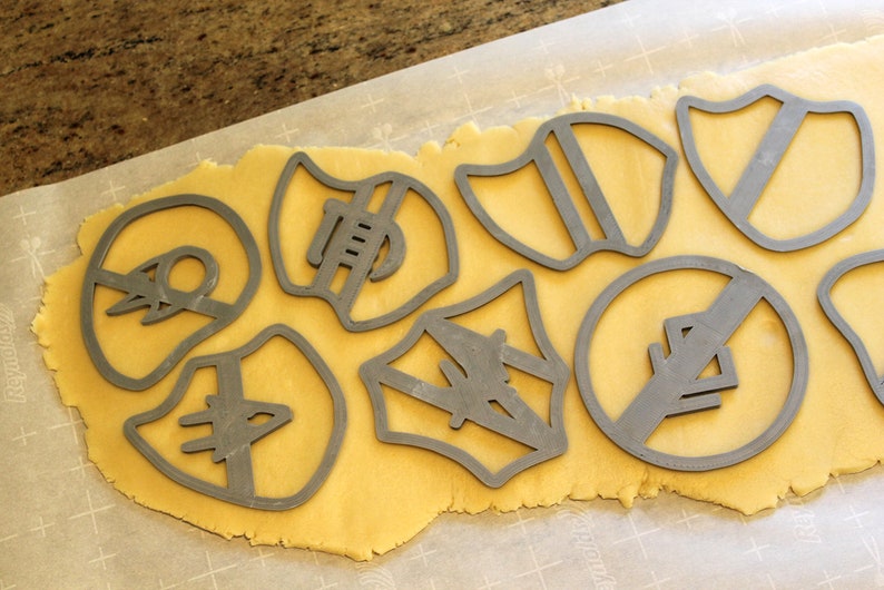 May include: A sheet of golden cookie dough with various cookie cutters pressed into it. The cutters are shaped like shields, circles, and other geometric shapes.