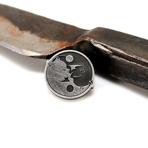 May include: A silver yin yang pin with a black cat and a white cat facing each other. The pin is sitting on top of a rusty knife.