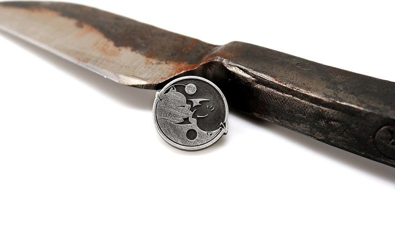 May include: A silver yin yang pin with a black cat and a white cat facing each other. The pin is sitting on top of a rusty knife.