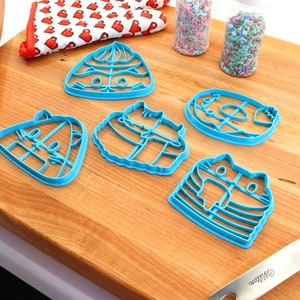 May include: Five blue plastic cookie cutters shaped like cats on a wooden chopping board. The cutters are arranged in a semi-circle with a rolling pin and a spatula in the background.
