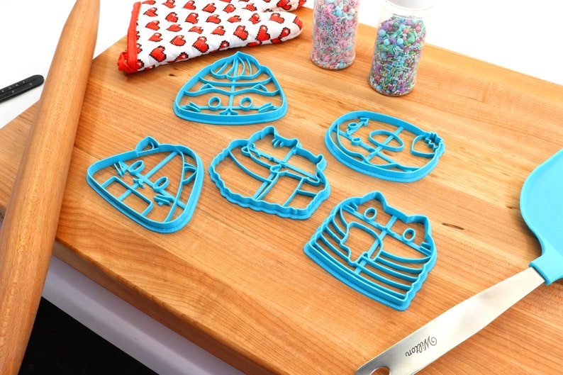 May include: Five blue plastic cookie cutters shaped like cats on a wooden chopping board. The cutters are arranged in a semi-circle with a rolling pin and a spatula in the background.