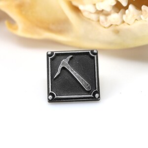K&ouml;nnte beinhalten: Ein schwarzer quadratischer Pin mit einem silbernen Pickel-Design. Der Pin hat einen strukturierten Rand und vier silberne Nieten.