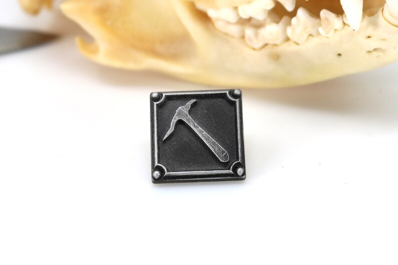 K&ouml;nnte beinhalten: Ein schwarzer quadratischer Pin mit einem silbernen Pickel-Design. Der Pin hat einen strukturierten Rand und vier silberne Nieten.