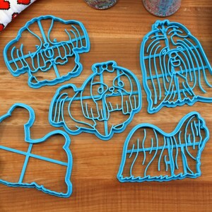 May include: Five blue plastic cookie cutters shaped like Shih Tzu dogs. The cutters are on a wooden surface.