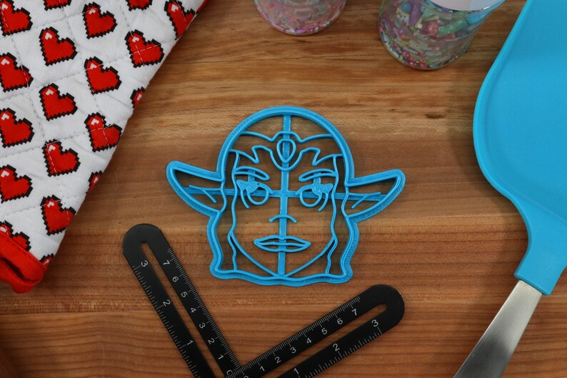 May include: Blue plastic cookie cutter shaped like a cartoon character with a crown and pointed ears. The character has a detailed face with a large nose and eyes.
