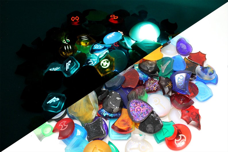 Puede incluir: Una colecci&oacute;n de piezas de juego de vidrio de colores, transl&uacute;cidas e iridiscentes con varios s&iacute;mbolos y dise&ntilde;os. Las piezas est&aacute;n dispuestas en una pila sobre una superficie blanca, con algunas de las piezas brillando en la oscuridad.