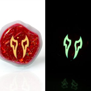 May include: A red, iridescent, resin-filled dice with a yellow glow-in-the-dark symbol of two flames. The dice is a polyhedron with many sides.