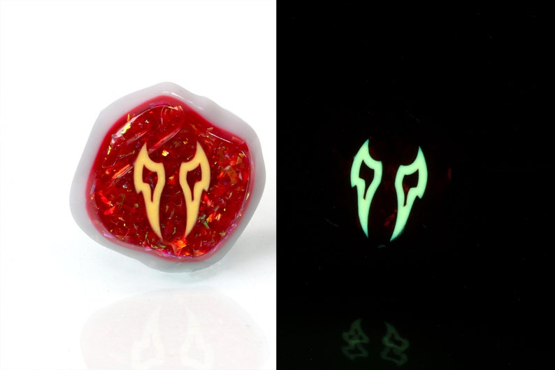 May include: A red, iridescent, resin-filled dice with a yellow glow-in-the-dark symbol of two flames. The dice is a polyhedron with many sides.