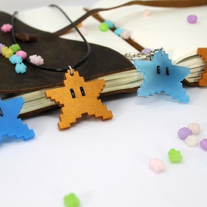 May include: Three star-shaped pendants with a pixelated design. The pendants are made of acrylic and come in blue, gold, and brown. The pendants are on a white surface with small, colorful candies scattered around them.