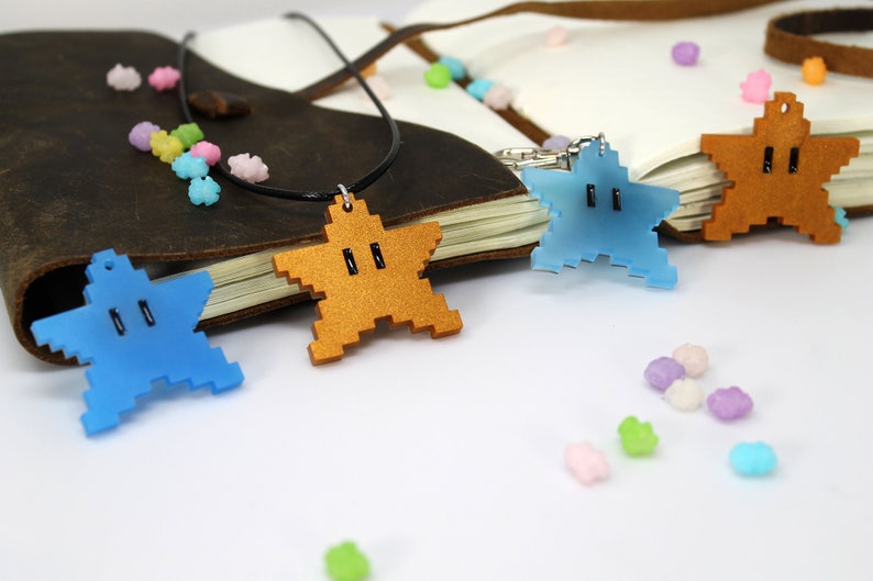 May include: Three star-shaped pendants with a pixelated design. The pendants are made of acrylic and come in blue, gold, and brown. The pendants are on a white surface with small, colorful candies scattered around them.