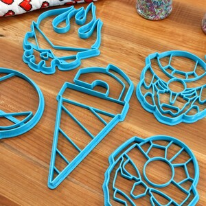 May include: Five blue plastic cookie cutters shaped like a skull, a flame, a cone, a donut, and a robot head.