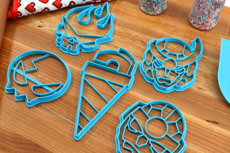 May include: Five blue plastic cookie cutters shaped like a skull, a flame, a cone, a donut, and a robot head.