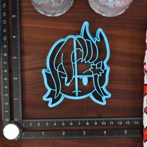 May include: A blue plastic cookie cutter shaped like a cartoon character with black hair and horns. The character has a surprised expression with one eye closed and the other open.