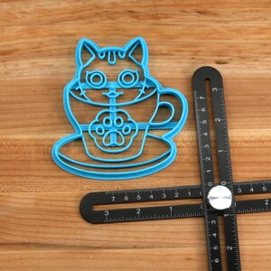 May include: Blue plastic cookie cutter shaped like a cat in a teacup. The cat has large eyes and a paw print on its chest. The teacup has a handle and a saucer.