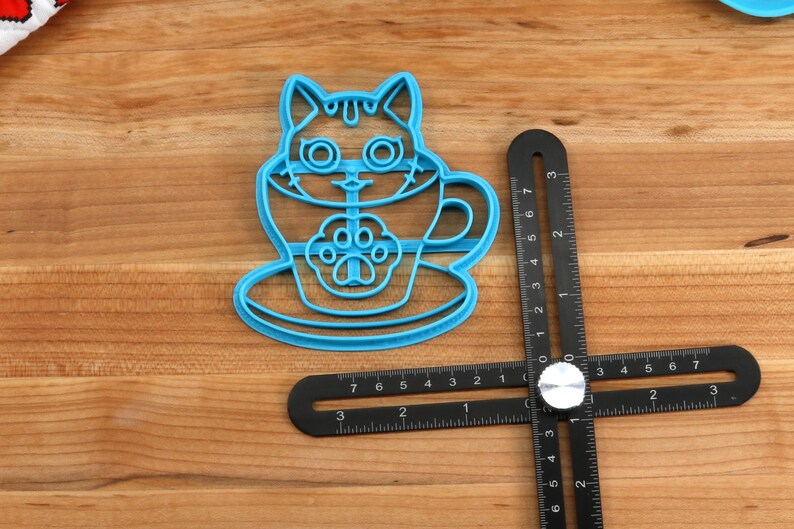 May include: Blue plastic cookie cutter shaped like a cat in a teacup. The cat has large eyes and a paw print on its chest. The teacup has a handle and a saucer.