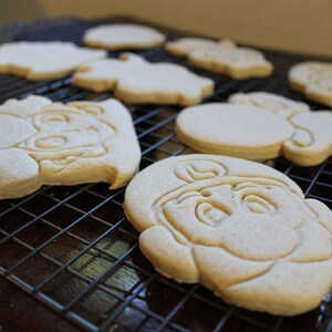 May include: A close-up of a cooling rack with several plain sugar cookies. The cookies are various shapes and sizes, including a round cookie with a face, a heart-shaped cookie, and a cookie shaped like a person.