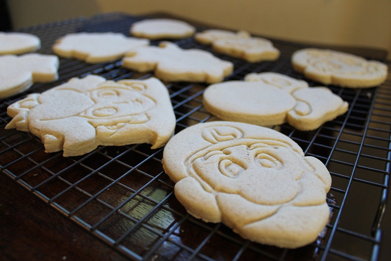 May include: A close-up of a cooling rack with several plain sugar cookies. The cookies are various shapes and sizes, including a round cookie with a face, a heart-shaped cookie, and a cookie shaped like a person.