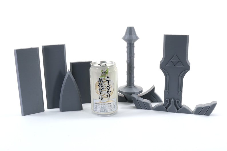 Puede incluir: Un conjunto de piezas de pl&aacute;stico gris para construir un modelo de espada. Las piezas incluyen la hoja, la empu&ntilde;adura y la guarda. Una lata de cerveza japonesa se encuentra en el centro de la imagen.