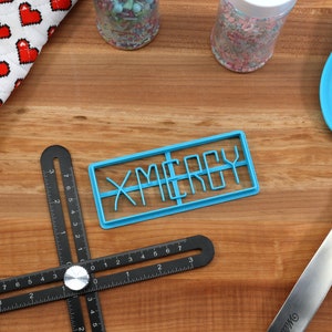 May include: A blue plastic cookie cutter shaped like the word "X-MERRY" on a wooden surface with a black ruler, a rolling pin, a blue spatula, and two jars of sprinkles.