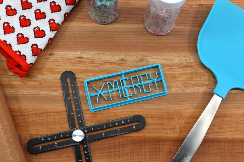 May include: A blue plastic cookie cutter shaped like the word "X-MERRY" on a wooden surface with a black ruler, a rolling pin, a blue spatula, and two jars of sprinkles.