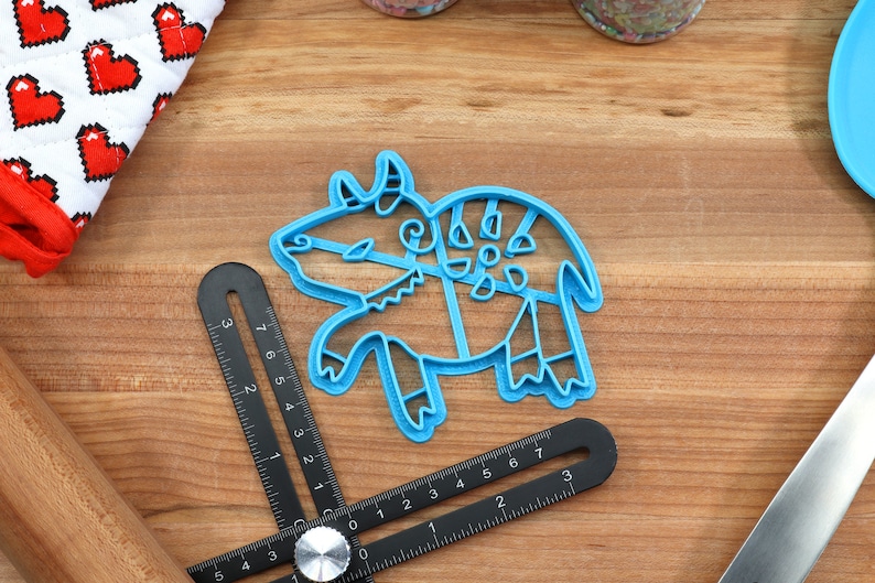 May include: Blue plastic cookie cutter shaped like a stylized bull with intricate details. The cutter is on a wooden surface.