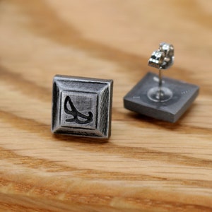 May include: A pair of silver square stud earrings with a black design. The earrings have a post back.