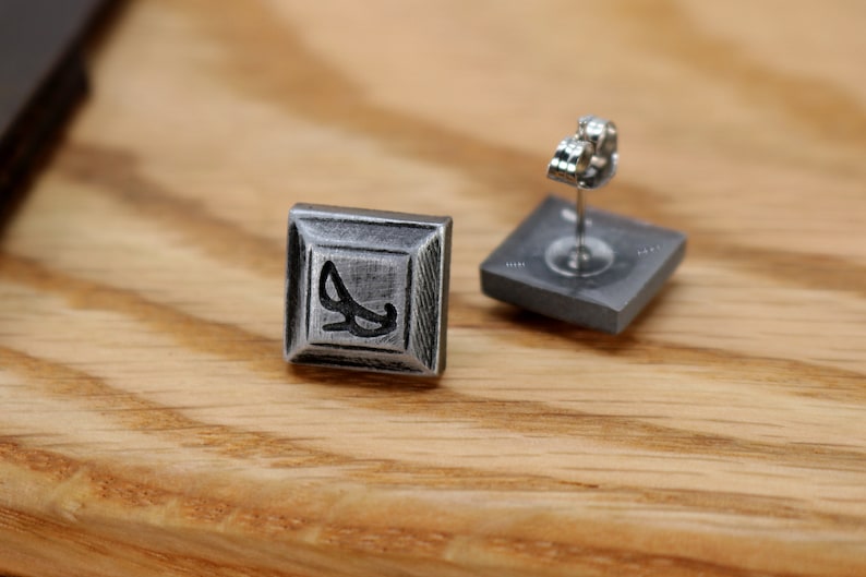 May include: A pair of silver square stud earrings with a black design. The earrings have a post back.