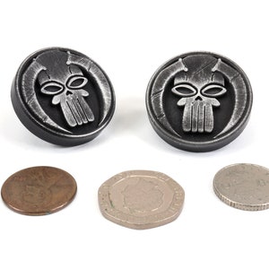 May include: Two silver-colored metal buttons with a skull design. The skull has a detailed design with horns and a textured surface. The buttons are about the size of a US penny.