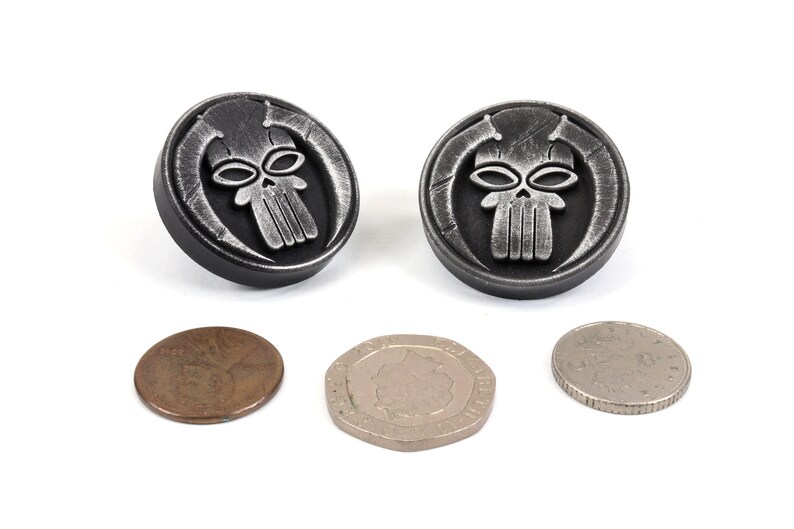 May include: Two silver-colored metal buttons with a skull design. The skull has a detailed design with horns and a textured surface. The buttons are about the size of a US penny.