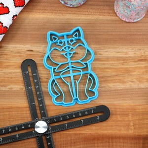 May include: A blue plastic cookie cutter shaped like a cartoon dog sitting with its paws crossed. The cutter is on a wooden surface with a ruler and other baking supplies.