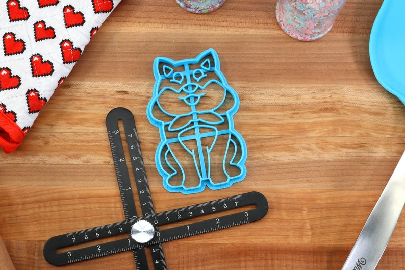 May include: A blue plastic cookie cutter shaped like a cartoon dog sitting with its paws crossed. The cutter is on a wooden surface with a ruler and other baking supplies.