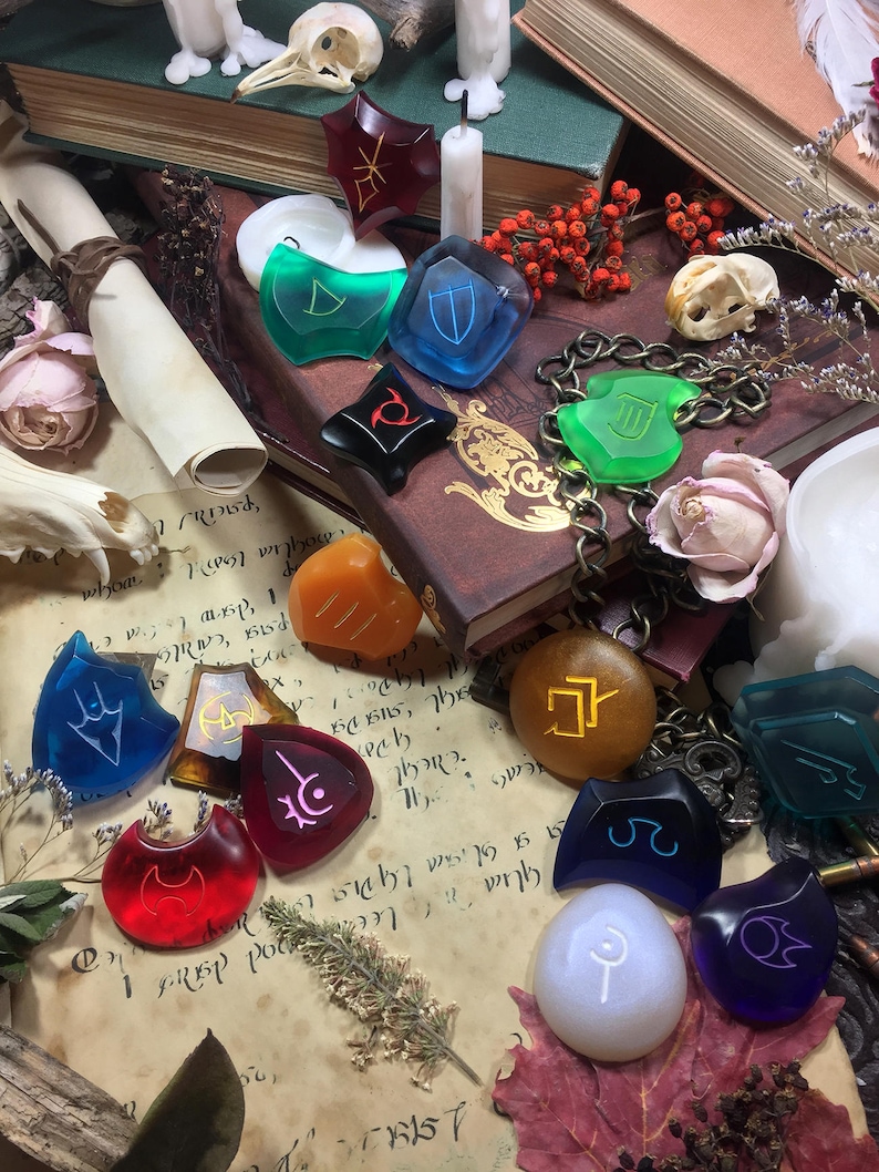 May include: A collection of colorful, translucent, and opaque glass and stone dice with various symbols and runes. The dice are arranged on a vintage book with a white candle, a skull, and a scroll.