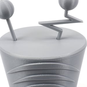 Pode incluir: Um modelo 3D impresso em cinza de um personagem de desenho animado estilizado com duas cabe&ccedil;as redondas e um bra&ccedil;o em forma de ziguezague.