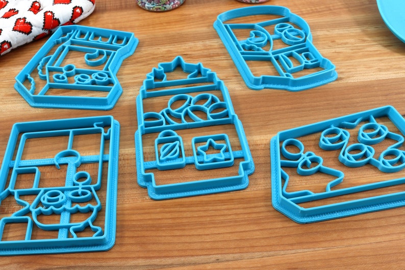 May include: Five blue plastic cookie cutters shaped like different vending machines. The cutters are on a wooden surface.