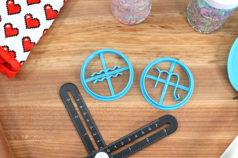 May include: Two blue plastic cookie cutters shaped like astrological signs. The cutter on the left is Aquarius and the cutter on the right is Virgo.