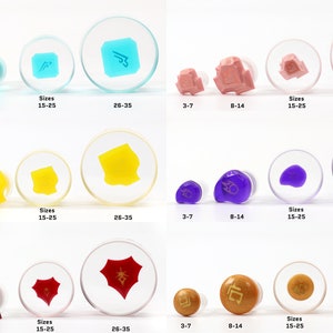 May include: Four sizes of clear acrylic ear gauges with different colored designs. The designs include a blue sword, a yellow shape, a purple blob, and a red Chinese character. The gauges are labeled with sizes 3-7, 8-14, 15-25, and 26-35.