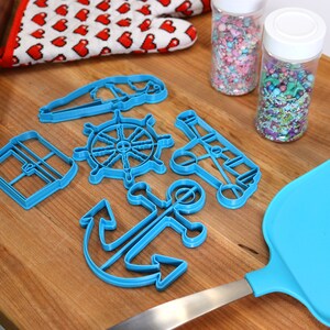 May include: A collection of blue nautical-themed cookie cutters, including an anchor, ship's wheel, and sailboat, arranged on a wooden surface. Also present are a blue spatula, a rolling pin, a heart-patterned oven mitt, and two jars of sprinkles.