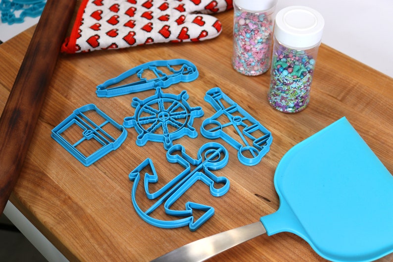 May include: A collection of blue nautical-themed cookie cutters, including an anchor, ship's wheel, and sailboat, arranged on a wooden surface. Also present are a blue spatula, a rolling pin, a heart-patterned oven mitt, and two jars of sprinkles.