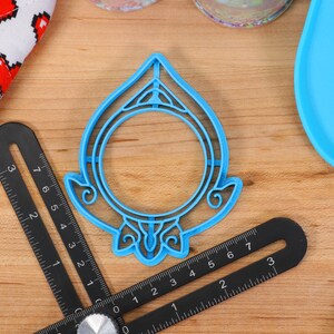 May include: A blue plastic cookie cutter shaped like a circle with a decorative design around the edge. The cutter is on a wooden surface.