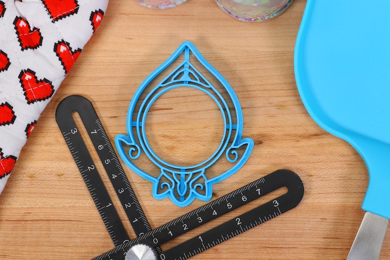 May include: A blue plastic cookie cutter shaped like a circle with a decorative design around the edge. The cutter is on a wooden surface.