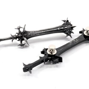 May include: Two black sword-shaped pins with intricate details. The pins have a silver metal clasp on the back.