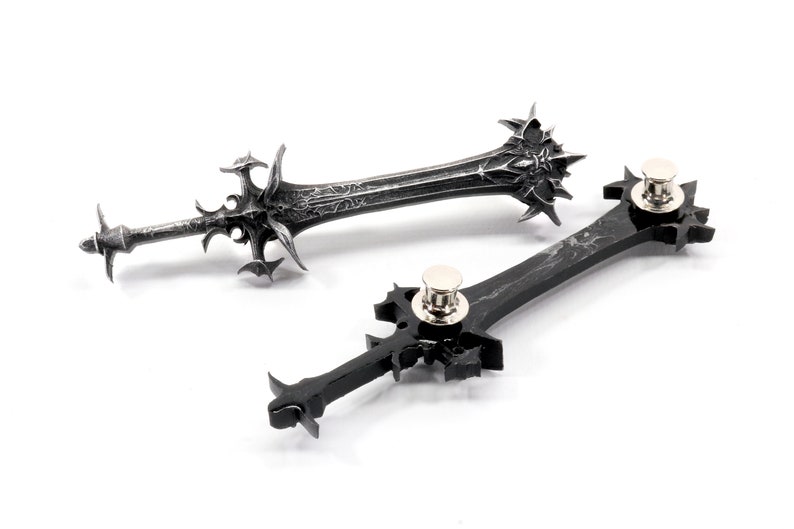 May include: Two black sword-shaped pins with intricate details. The pins have a silver metal clasp on the back.