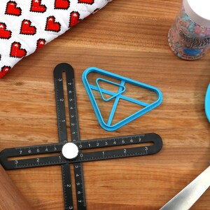May include: A blue plastic triangle-shaped mold for making sushi, a black ruler with a rotating dial, a wooden rolling pin, a blue silicone spatula, and a jar of colorful sprinkles.