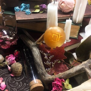 Puede incluir: Una imagen de bodeg&oacute;n con un orbe dorado con un s&iacute;mbolo, rodeado de velas, libros, flores secas y una botella de vidrio con un tap&oacute;n de corcho.