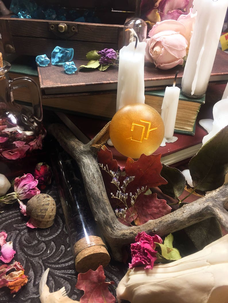 Puede incluir: Una imagen de bodeg&oacute;n con un orbe dorado con un s&iacute;mbolo, rodeado de velas, libros, flores secas y una botella de vidrio con un tap&oacute;n de corcho.