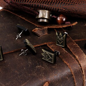 May include: A collection of stud earrings with a crown and heart design. The earrings are in black and bronze tones, with silver posts. They are displayed on a brown leather surface, alongside a leather bag.