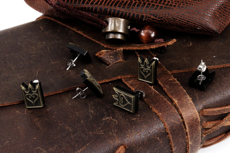 May include: A collection of stud earrings with a crown and heart design. The earrings are in black and bronze tones, with silver posts. They are displayed on a brown leather surface, alongside a leather bag.